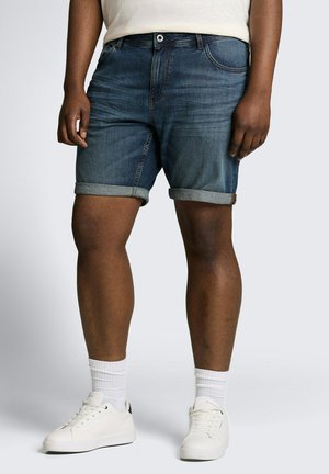 Person, die dunkelblaue hochgekrempelte Jeansshorts, weiße Kniestrümpfe und weiße flache Sneakers trägt und vor einem schlichten Hintergrund steht.