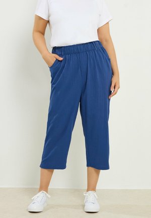 Persona vistiendo pantalones cortos sueltos azules con cintura elástica y bolsillos, combinados con zapatillas blancas y una camiseta blanca de manga corta.