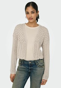 Cardigan lavorato a maglia beige chiaro con apertura anteriore, caratterizzato da un motivo in pizzo, maniche lunghe e orlo a scallop, indossato sopra una camicia chiara e jeans.