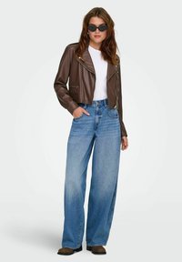 Jachetă de biker din piele maro, tricou alb și blugi largi albaștri. Ochelari de soare și cizme scurte maro completează ținuta.