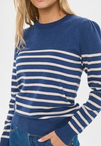 Modrý pruhovaný sveter s krémovými horizontálnymi pruhmi, rebrovanými manžetami a lemom, okrúhlym výstrihom a mäkkou pletenou textúrou. Priliehavý strih so slabými vyp puff rukávmi.