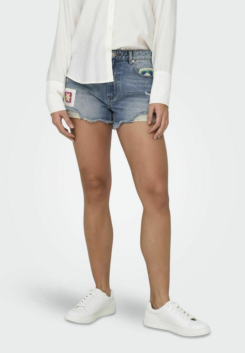 Pantalones cortos de mezclilla con un dobladillo desgastado, que presentan parches de bordado multicolor en la parte delantera. Combinados con una camisa blanca de botones y zapatillas blancas.