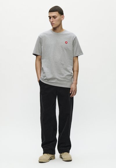 T-shirt en coton gris à manches courtes, arborant un petit logo rouge sur la poitrine ; assorti à un pantalon en velours côtelé noir et des chaussures beiges.