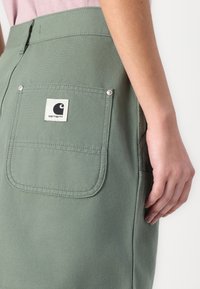 Fustă din bumbac verde măslin, cu un buzunar mare pe spate, dotat cu un eticheta albă, cusături evidențiate și accente hardware vizibile.