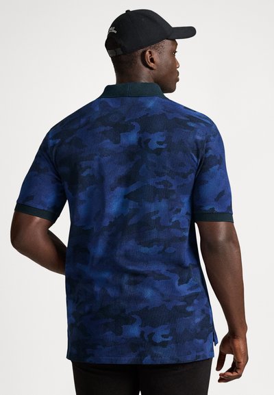 Mężczyzna ubrany w niebiesko-czarną polówkę w kamuflażu i czarną czapkę, widziany od tyłu na jednolitym tle.