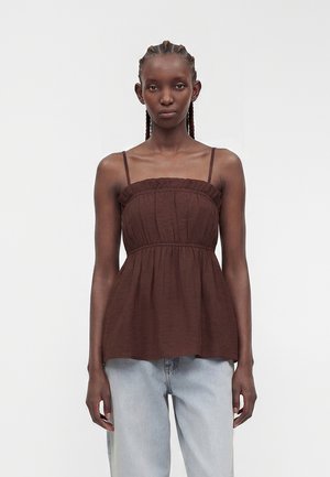 Femme aux cheveux tressés portant un haut marron sans manches froncé avec des bretelles fines et un jean bleu clair, debout devant un fond uni.