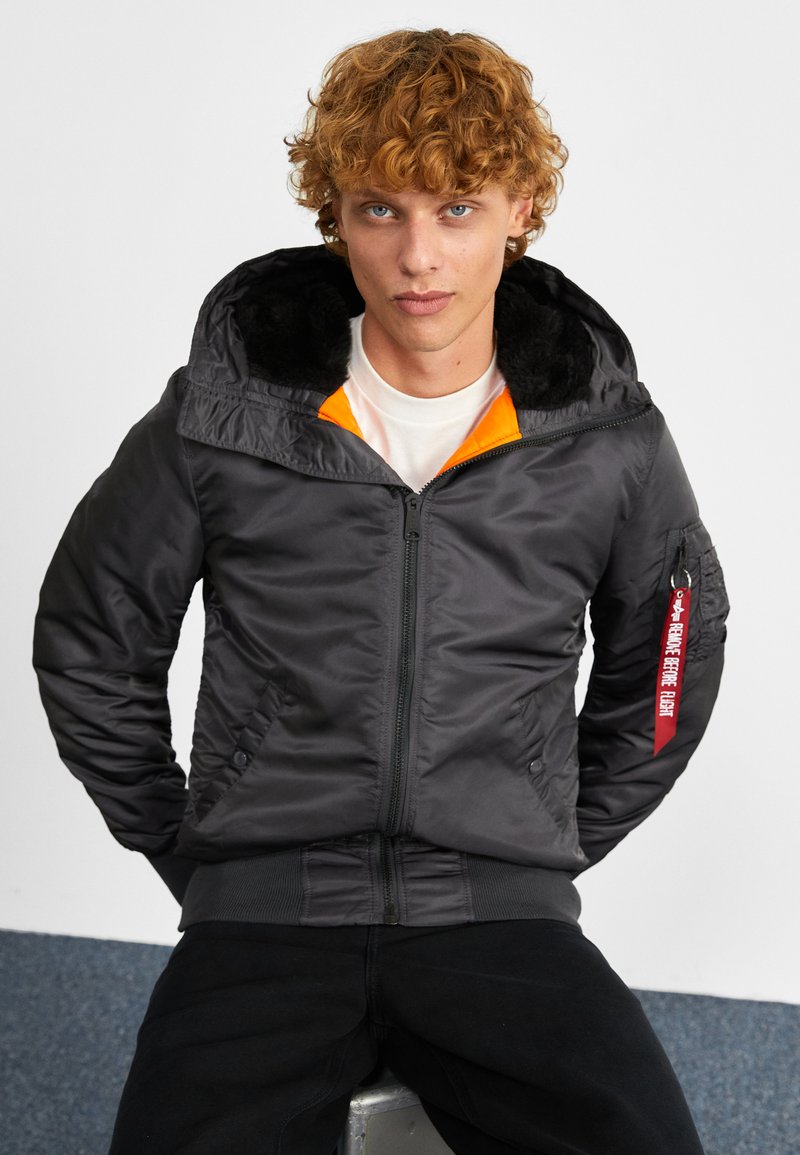 Black bomber jacket with faux fur lining, orange collar, zippered front, side pockets, and red label detail on sleeve. Smooth, padded texture.