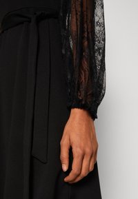Main droite à côté d'une robe noire avec manches transparentes en dentelle et ceinture en tissu nouée à la taille, sur un fond clair uni.