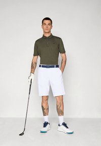 Πράσινο πόλο πουκάμισο με τρία κουμπιά, λευκά σορτς, μπλε ζώνη, λευκά παπούτσια γκολφ με ναυτικό μπλε λεπτομέρειες, κρατώντας ένα μπαστούνι γκολφ. Ορατά τατουάζ στο χέρι.