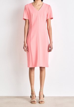 Personne portant une robe rose à manches courtes, longueur genoux, avec un col en V et des sandales beiges à lacets, debout sur un fond uni.