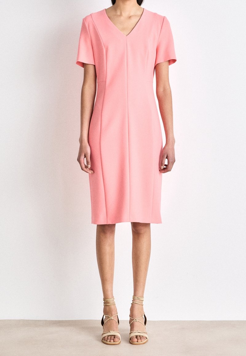 Personne portant une robe rose à manches courtes, longueur genoux, avec un col en V et des sandales beiges à lacets, debout sur un fond uni.