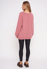 Rosa, flauschiger Strickpullover mit lockerer Passform und überschnittenen Schultern, kombiniert mit schwarzen Leggings. Die Textur ist weich und leicht.