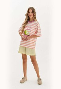 Žena stojí oblečená v ružovo-bielom pruhovanom tričku s grafikou jablka, svetložltých šortkách a béžových slip-on topánkach.