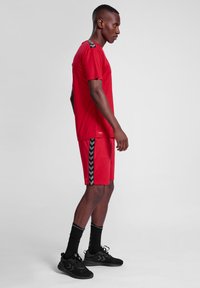 Camiseta deportiva roja y pantalones cortos, con acentos negros de espiga en los lados. Llevado con zapatillas deportivas negras y calcetines.