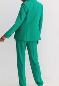 Blazer verde su misura e pantaloni coordinati realizzati in tessuto liscio. Il blazer presenta un colletto a punta e dettagli con bottoni sulle maniche.