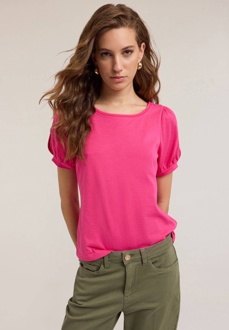 Top scurt roz cu mânecă cu umbrelă și guler rotund, asortat cu pantaloni verzi măsliniu cu închidere frontală cu nasture.