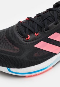 Zwarte sportieve schoen met een gestructureerde mesh bovenwerk, voorzien van roze accenten, zwarte veters en een witte tussenzool met blauwe en roze details.