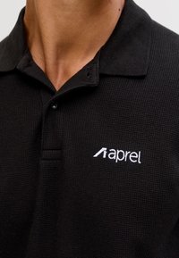 Close-up van een persoon die een zwart, getextureerd poloshirt draagt met een klein wit "aprel" logo op de linkerborst.