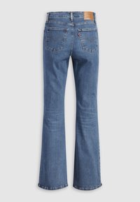 Jean en denim bleu avec jambes évasées, deux poches arrière, patch de marque en cuir sur la ceinture, et une petite étiquette en tissu rouge sur la poche droite.