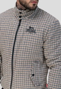 Veste à carreaux dans des tons de marron et de noir. Présente un col montant, un zip à l'avant et une poche. Logo sur la poitrine. Tissu texturé.