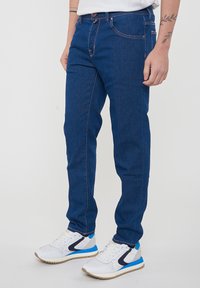 Jeans in denim blu dalla vestibilità slim, con cuciture a contrasto rosa, cinque tasche e chiusura a bottoni, abbinati a sneakers bianche e blu.