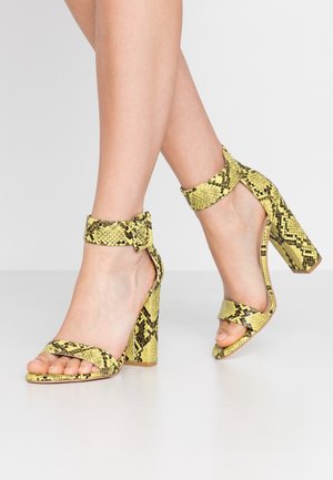 Sandales à talons hauts avec un motif en peau de serpent jaune et noir, dotées de brides à la cheville et de talons épais. Design à bout ouvert avec une finition texturée.