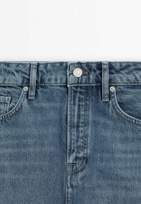 Denimjeans i blått med midjeband som visar knapp, bältesöglor, framfickor och sömnadsdetaljer på en vit bakgrund.