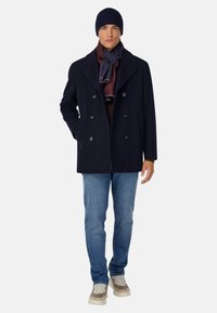 Marineblauer, doppelt geknöpfter Mantel aus Wollmischung mit großen Knöpfen. Kombiniert mit einem burgunderfarbenen und blauen Schal, Jeans und beigen Sneakers. Strickmütze in Marineblau.