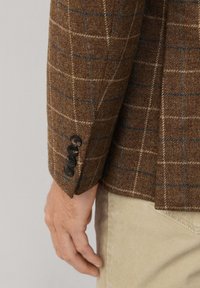 Braun kariertes Blazer aus strukturiertem Wollstoff mit blauen und beigen Linien. Vier schwarze Knöpfe am Ärmel, taillierte Passform.