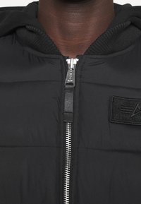 Nærbillede af en person iført en sort dunjakke med en sølvfarvet lynlås og et sort mærke med et logo på brystet.