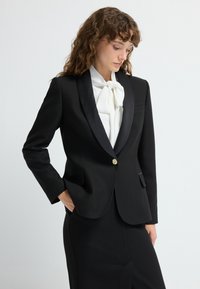 Černý konfekční blazer se saténovými klopami, zlatým detailem na knoflíku a dvěma předními kapsami, v kombinaci s bílou halenkou s mašlí. Hladká textilie.