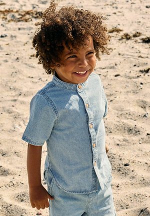 Enfant souriant aux cheveux bouclés portant une chemise rayée bleu clair à manches courtes et un short assorti, debout sur un sol sablonneux.