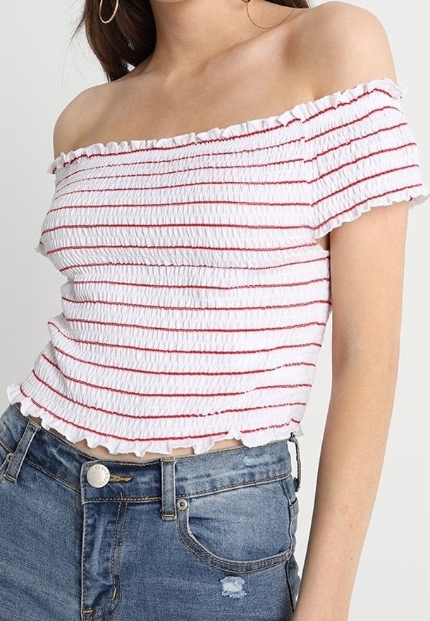 Femme portant un top blanc épaules dénudées avec des rayures rouges et des bords volantés, associé à un jean en denim bleu.