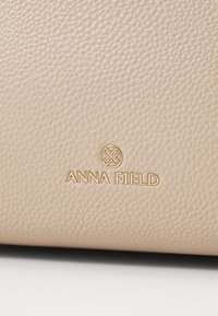 Surface en cuir beige texturé avec un petit logo circulaire doré et le nom de la marque Anna Field embossé en dessous.