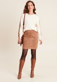 Frau mit einem weißen, flauschigen Pullover, einem braunen Wildlederrock, schwarzen Strumpfhosen, kniehohen braunen Stiefeln und einer braunen Umhängetasche.