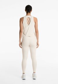 Beige atletisk linne med detaljerad crisscross-baksida, i kombination med åtsittande beige leggings och vita sneakers. Slät textur och minimalistisk design.