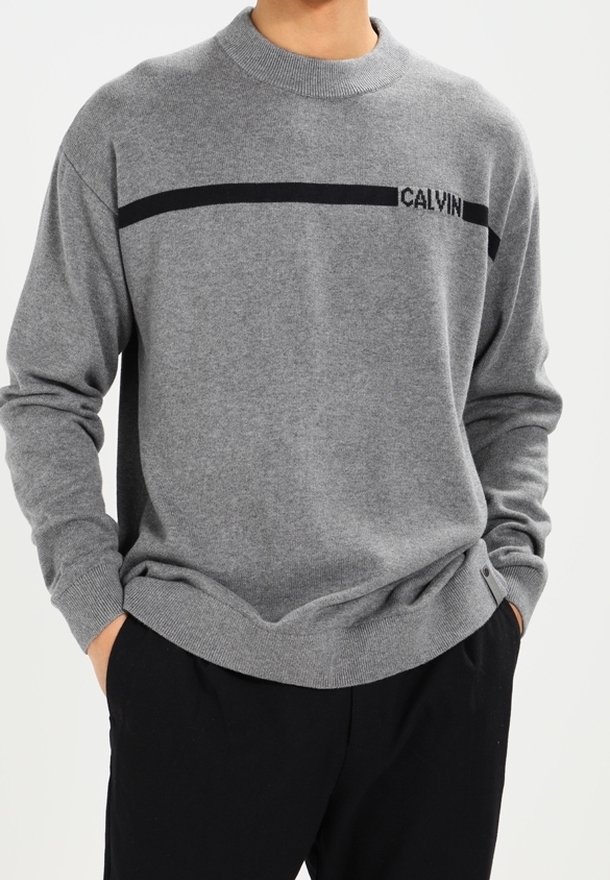 Personne portant un pull gris avec une bande horizontale noire et un texte partiel "CALVIN", mains dans les poches, assorti à un pantalon noir.