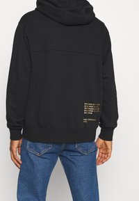 Svart hoodie med huva och ribbade muddar. Har guldig text tryckt på nedre delen av ryggen. Kombinerad med blå denimjeans, som visar en avslappnad stil.