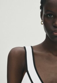 Weißes Tanktop mit schwarzem Saum, aus strukturiertem Stoff. Sichtbares goldenes Ohrring aus einem Hoop. Dunkle Hautfarbe. Nahaufnahme von Schulter und Gesicht.