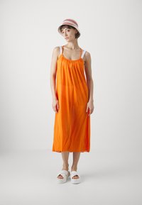 Vestido maxi sin mangas de color naranja con escote fruncido, tirantes blancos y una textura ligera. Combinado con sandalias de plataforma blancas y un sombrero a rayas.