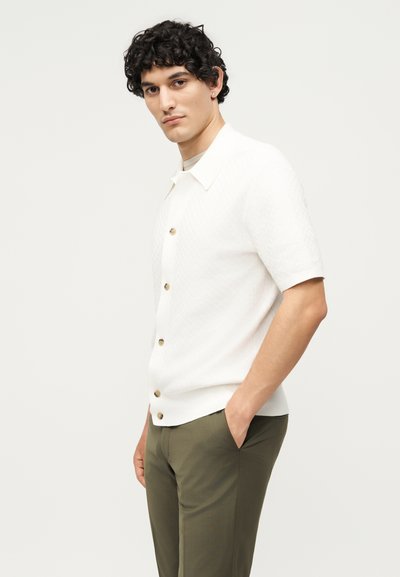 Νέος άνδρας με σγουρά σκούρα μαλλιά φοράει ένα άσπρο κοντομάνικο πουκάμισο με κουμπιά και ελιά πράσινο παντελόνι, ποζάροντας με το ένα χέρι στην τσέπη.