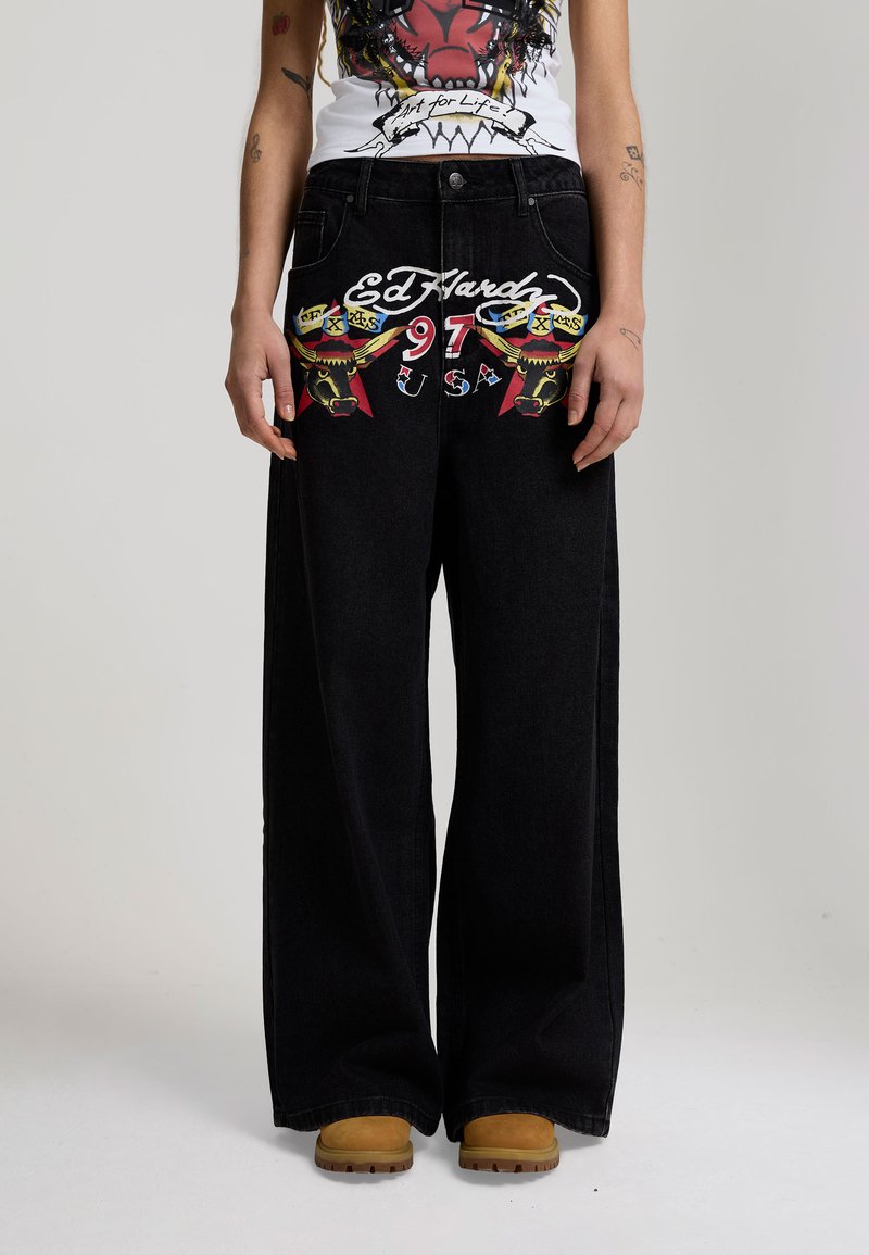Zwarte jeans met wijde pijpen en kleurrijke stiergrafieken, de teksten "Ed Hardy," "97," en "USA" aan de voorkant, gedragen met beige laarzen en een grafisch shirt.