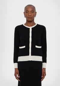 Black cardigan with cream accents, featuring a round neck, button closure, two front pockets, and ribbed cuffs and hem. Knitted texture.