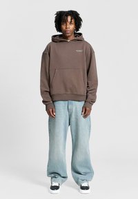 Brown oversized hoodie with front pocket and logo detail, paired with light blue wide-leg jeans and black and white sneakers.