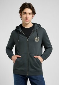 Felpa zip verde scuro con cappuccio regolabile, tasche frontali e un logo discreto sul lato sinistro del petto, realizzata in tessuto morbido e testurizzato.
