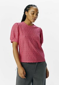 Haut à manches bouffantes rose avec un motif de perforation en diamant, tissu texturé, encolure ronde, associé à un pantalon gris.