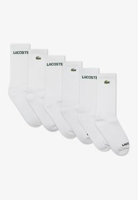 Lot de six chaussettes blanches en coton avec texture côtelée, arborant le logo Lacoste vert brodé en haut et un lettrage noir sur la semelle.