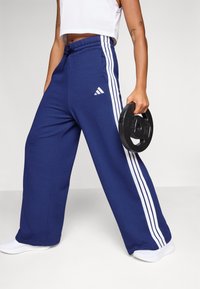 Mornarice plave trenirke od mekanog materijala, sa bijelim bočnim trakama i Adidas logotipom. Širokog kroja s podesivim pojasom.