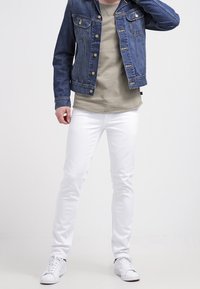 Jeansjack met blauwe wassing, twee borstzakken, beige t-shirt, witte slim-fit jeans en witte sneakers, wat een casual outfit toont.