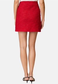 Mini-jupe rouge texturée à la coupe ajustée, avec un ourlet brut. Des talons noirs à fines lanières complètent la tenue, la peau lisse étant visible.